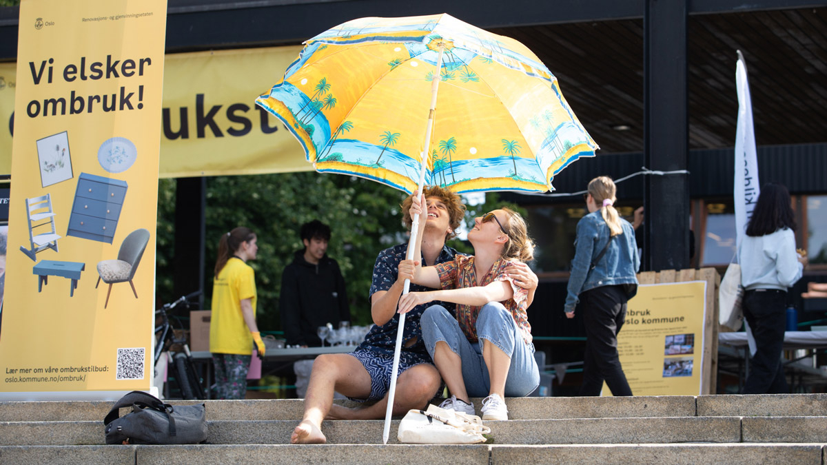 Two students sitting under a yellow umbrella at an event at a square