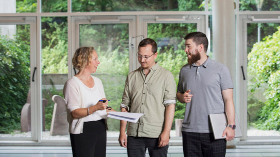Three researchers discussing a document, standing in front of a large window
