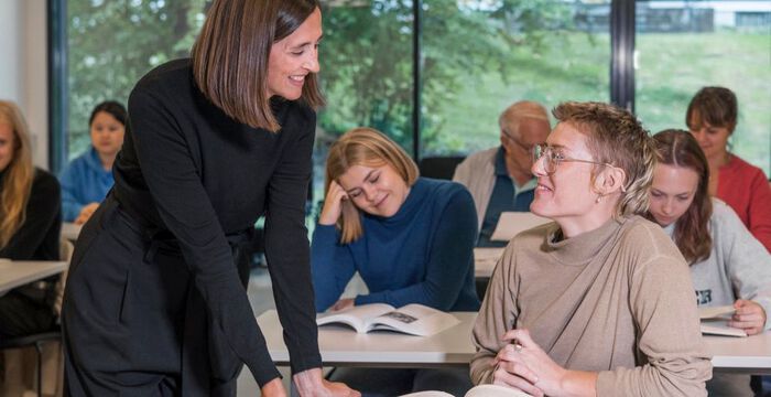 Undervisningsrom med studenter. Kvinnelig l?rer st?r ved pulten til en elev og de smiler til hverandre. 