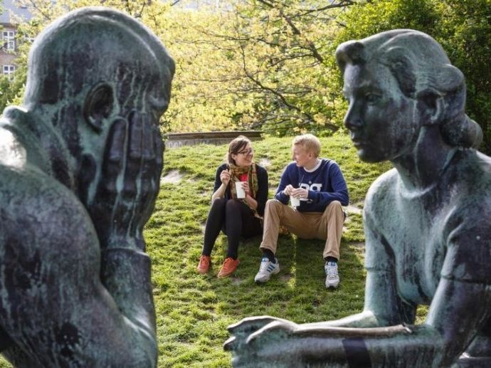 To mennesker sitter p? en gressbakke og snakker sammen. I forgrunnen ser vi to statuer som snakker sammen.
