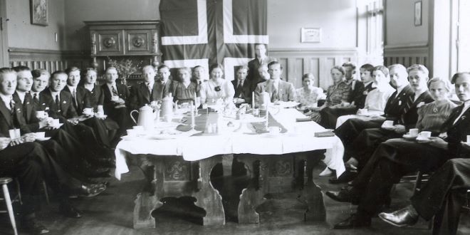 Svart-hvitt fotografi. Menn og kvinner sitter p? stoler langs rommets vegger, med et stort bord i midten. Det norske flagg henger p? bakveggen.