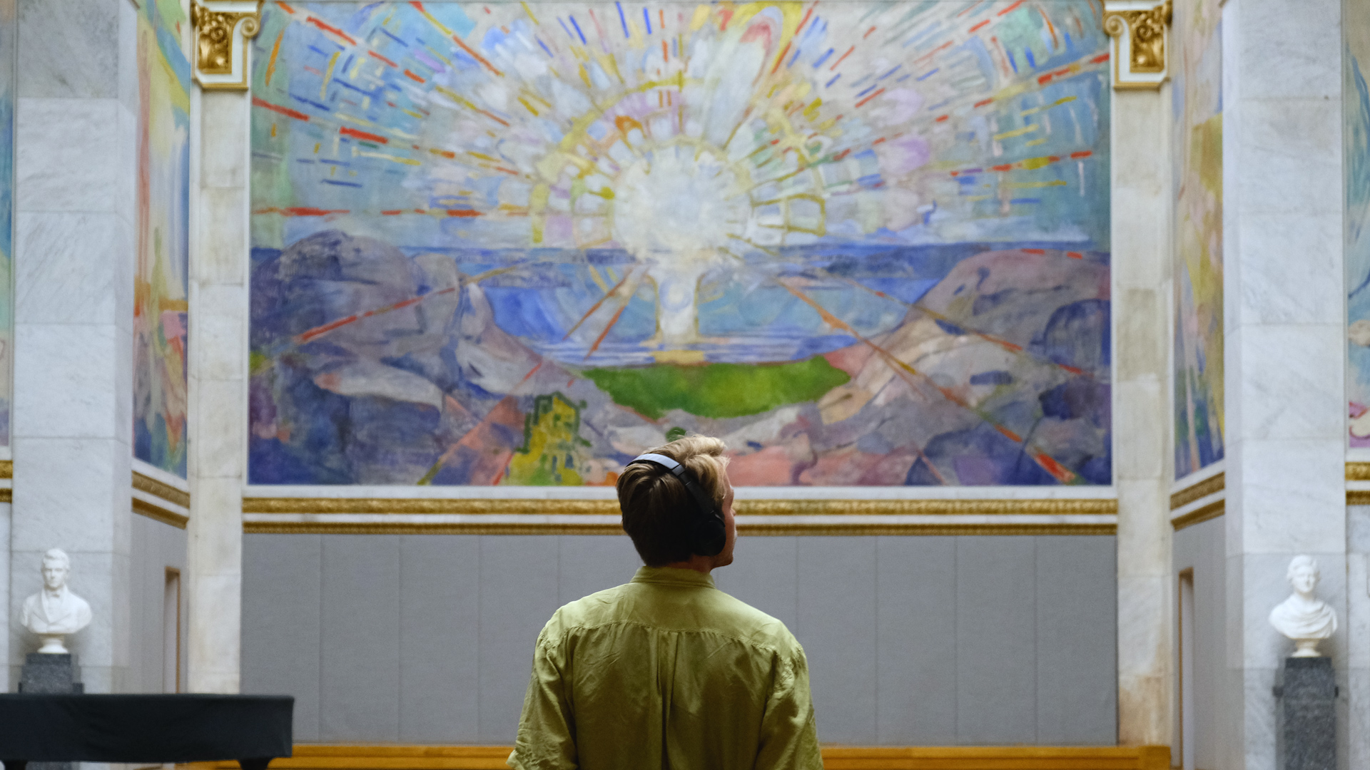 Mann i gr?nn skjorte forgrunnen, som har p? seg headset. I bakgrunnen ser vi Edvard Munchs maleri "Solen", som henger i Universitets aula.