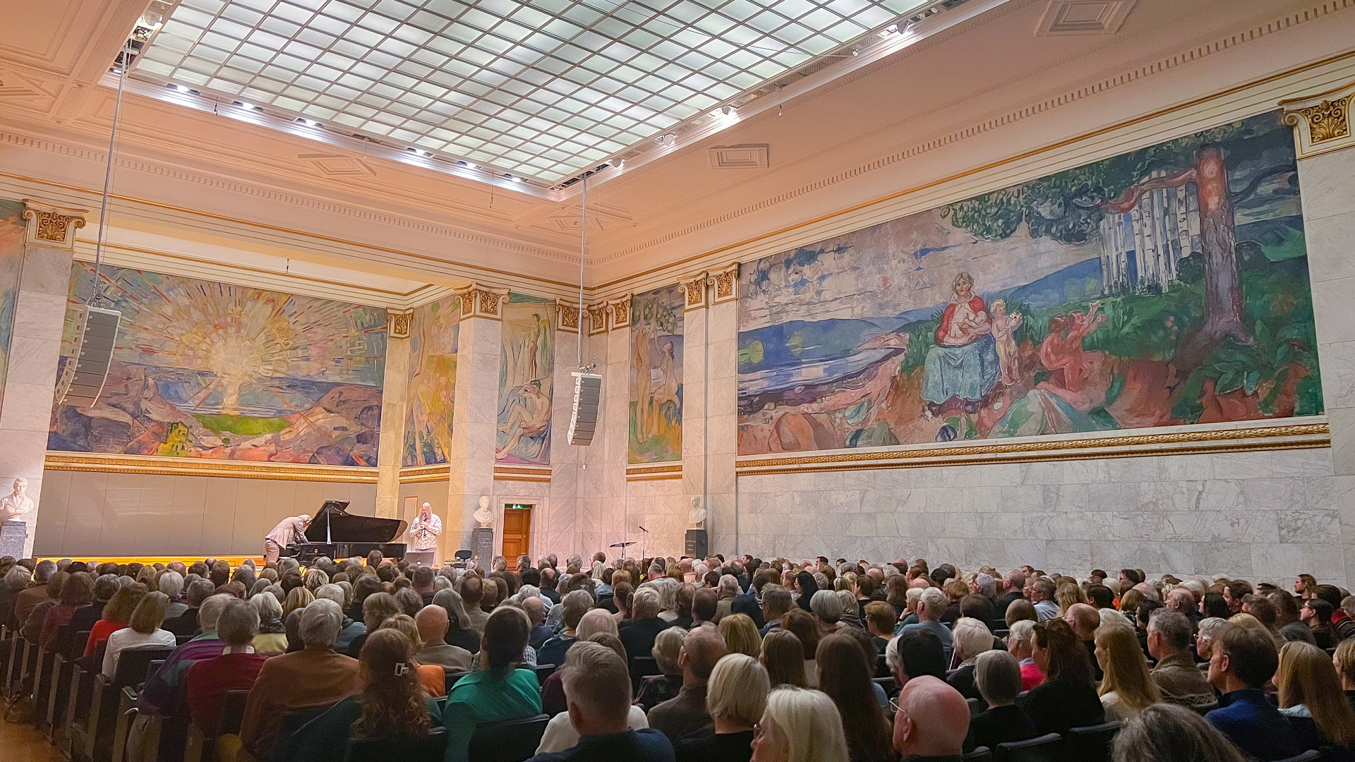 Universitetets aula er fullsatt, med publikum vendt mot scenen der en pianist og en gitarist spiller. I bakgrunnen ser vi Munchs malerier "Alma Mater" og "Solen".