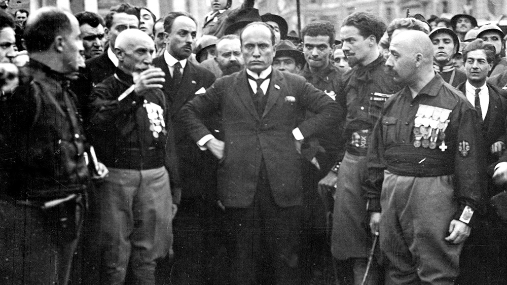 Svart-hvitt bilde av en dresskledd Benito Mussolini blant en gruppe menn i svarte skjorter.
