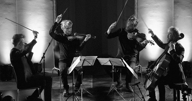 Photo of the Danish String Quartet, four string musicians seated, playing, lifting their bows at the same time