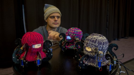 Man standing behind a table with three Dr. Squiggles-robots. The robots look like octopuses with knitted balaclavas.