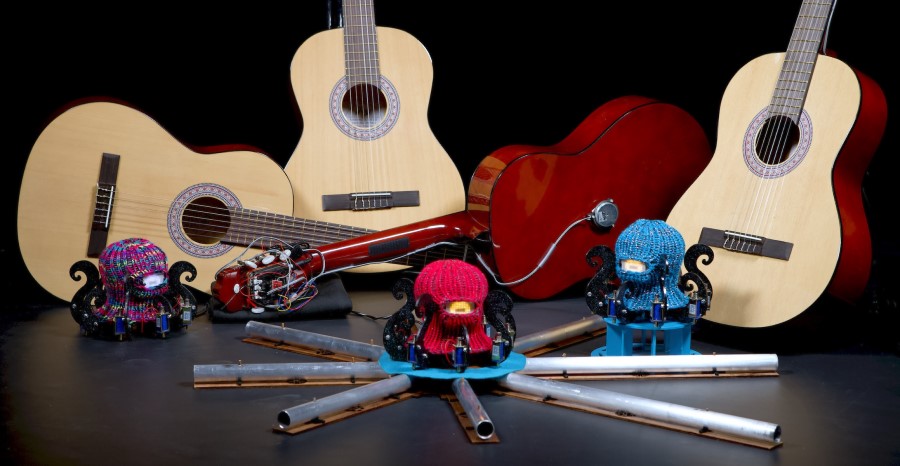 Tre Dr. Squiggles-roboter med gitarer i bakgrunnen. Robotene ser ut som blekksputer med strikkede finlandshetter.