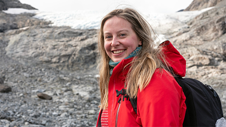 Kvinnelig student smiler mot kamera i turkl?r foran et sn?kledd fjell